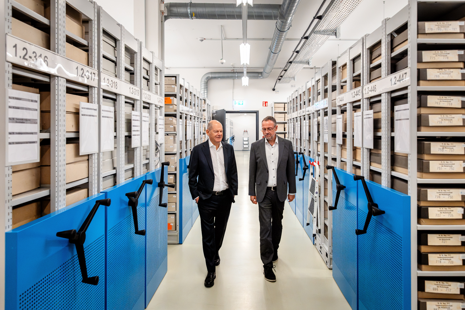 Olaf Scholz und Mario Glauert gehen durch einen Gang mit blauen Rollregalanlagen voller Aktenkartons