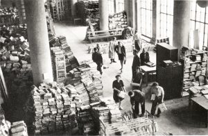 Ein Schwarz-Weiß-Foto zeigt Mitarbeitende in einer großen Halle, die Aktenberge sortieren